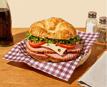 Golden croissant sandwich with ham, cheese, lettuce, and tomato on a checkered napkin beside a soda bottle. Text reads "Lunch."