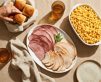 Overhead view of sliced ham and turkey platter, macaroni and cheese, dinner rolls, and iced tea on linen. Text reads "Meals."